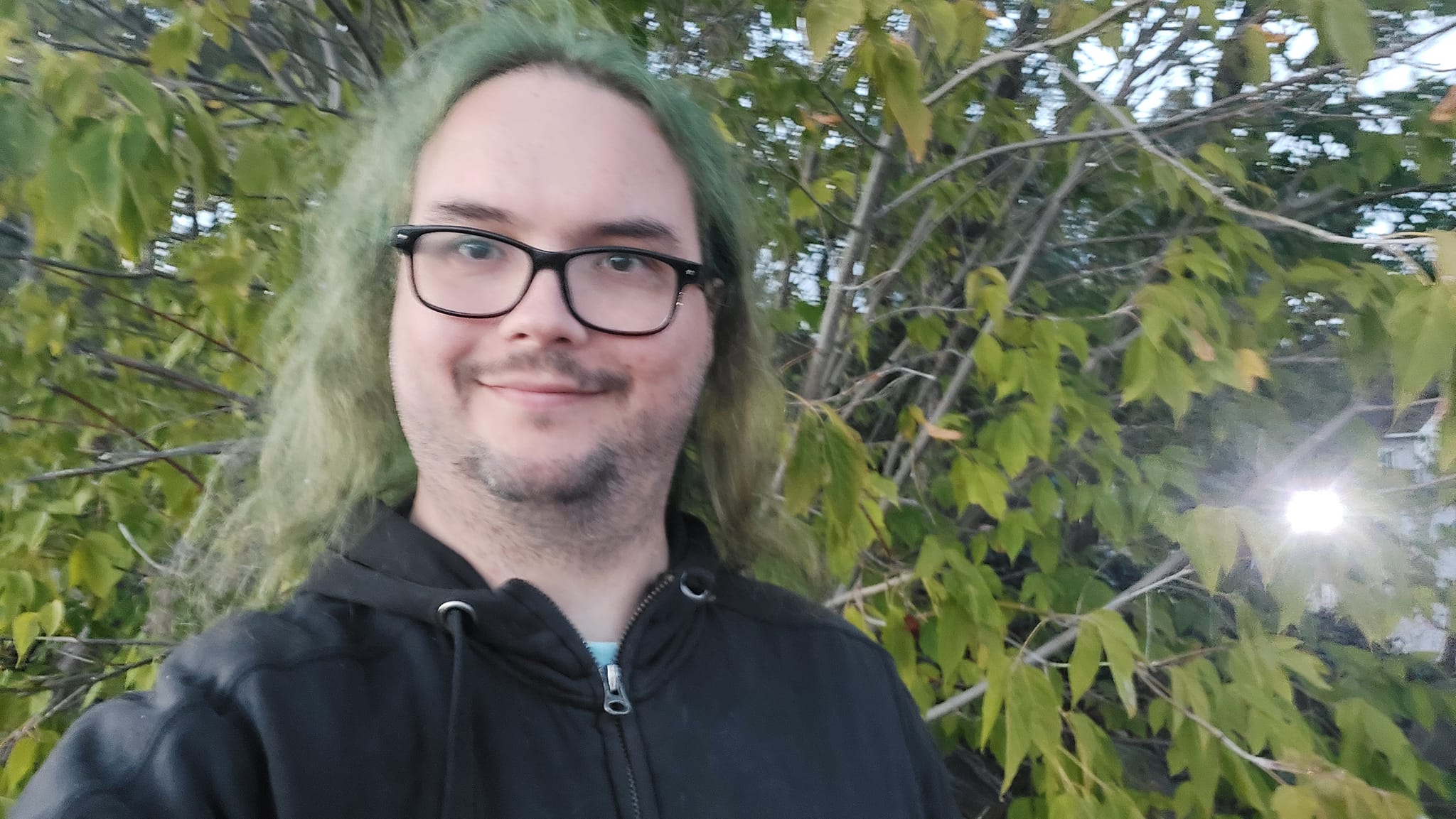 A man with long green hair and no beard stands outside in front of a tree with leaves